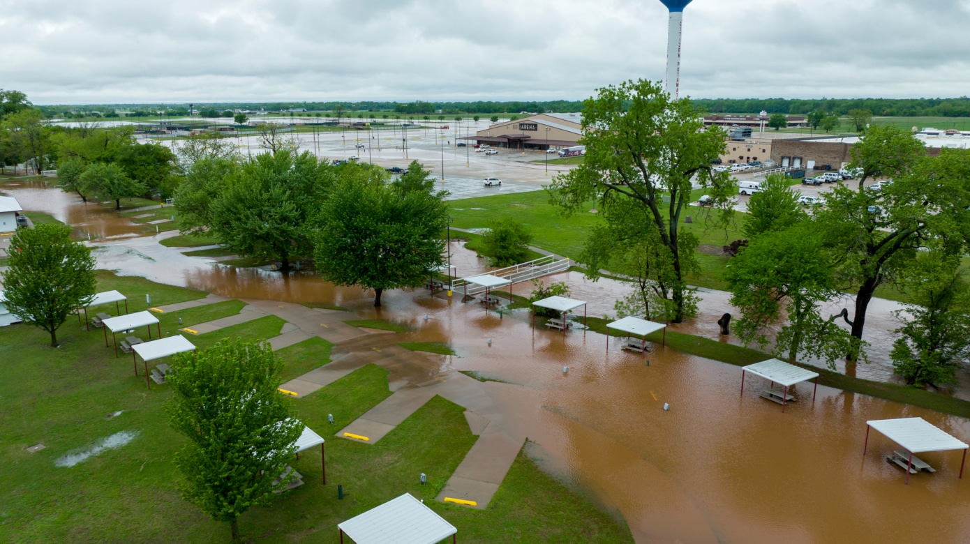 An aerial photo of camping sites on CPN's Festival grounds flooded with muddy brown water. The CPN water tower and FireLake Arena are visible in the background. The green trees emerging from the flood waters make a striking color contrast with the red-brown water.