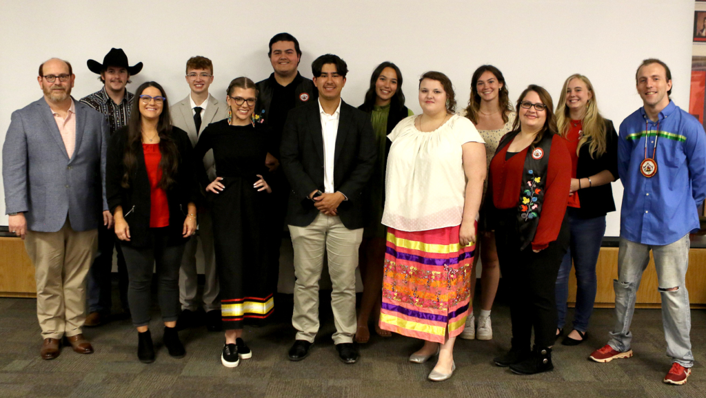 A group of students across a range of ages and genders, some wearing blazers, some wearing ribbon skirts and ribbon shirts, and one wearing a woodlands floral graduation stole, stand together for a photograph at the 2023 CPN Department of Education graduation celebration.