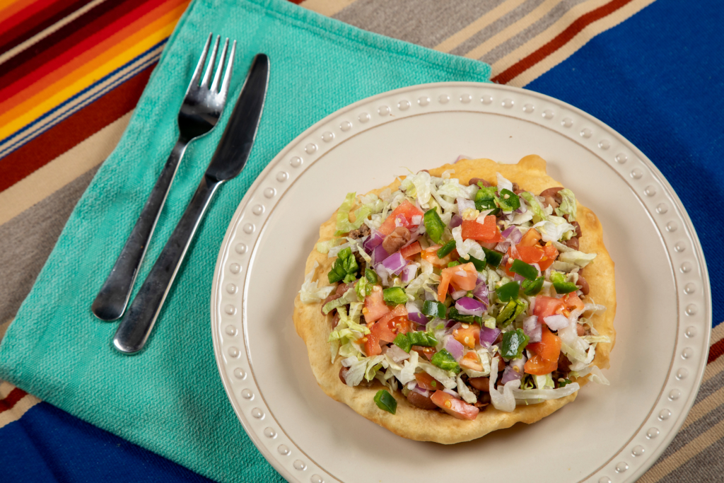 A piece of fry bread on a white plate piled high with lettuce, peppers, tomatoes, beans, meat, and more.