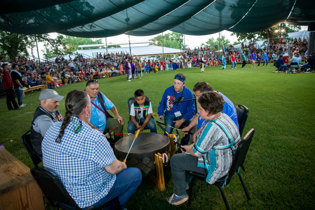Several Citizen Potawatomi men sit around the drum in the dance arena at the 2022 Familly REunion Festival. Festival participants can be seen in the background. 