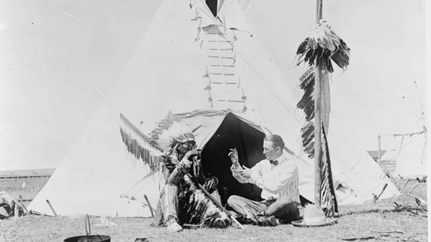 Two people seated in front of a teepee communicate in Plains Indian Sign Language.