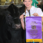A smiling Helen Spears, with her hair braided neatly to one side, stands with her calf Fancy, a black heifer she bought one year ago. She holds her purple award banner in her left hand.