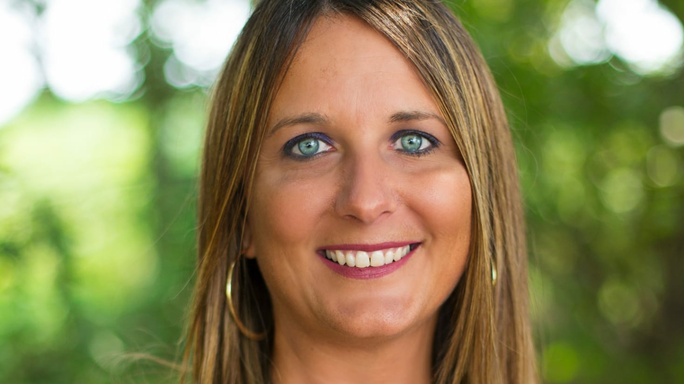 Headshot of Self-Governance director Kasie Nichols. She stands in front of green foliage and wears a blue patterned blouse. Her brown hair falls past her shoulders and her blue eyes sparkle.