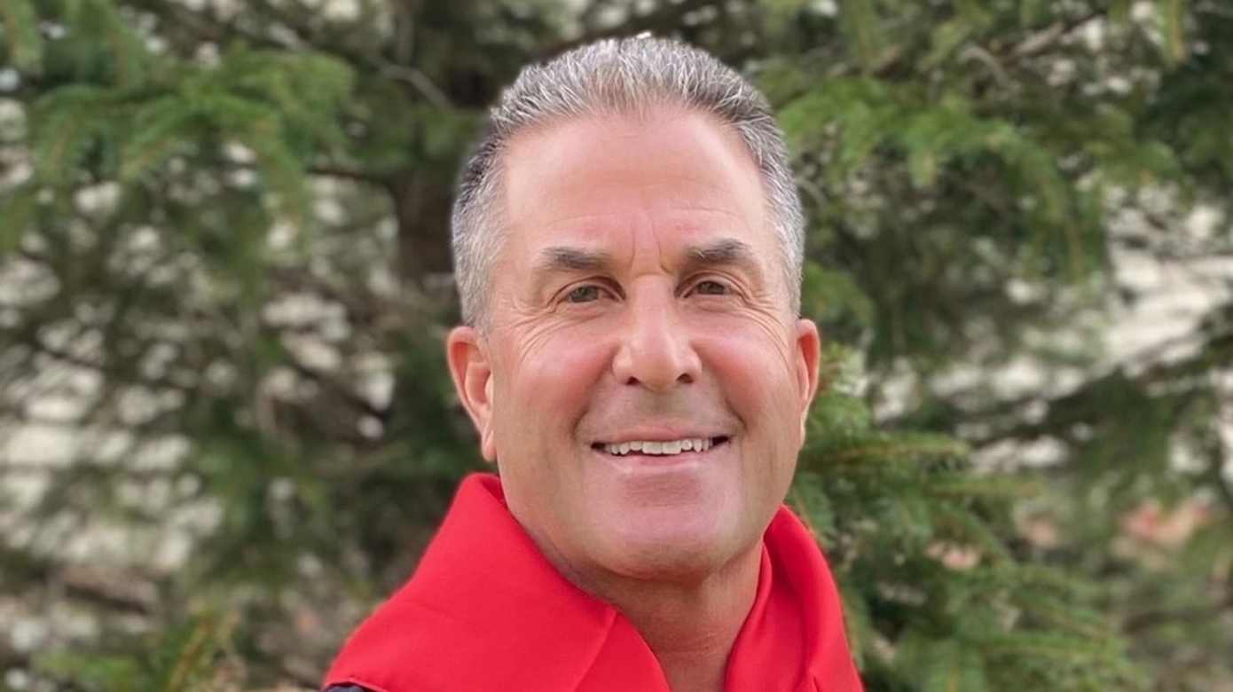 Photo of Tribal member Kevin Roberts in a ribbon shirt and red scarf standing in front of a pine tree.