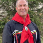 Photo of Tribal member Kevin Roberts in a ribbon shirt and red scarf standing in front of a pine tree.