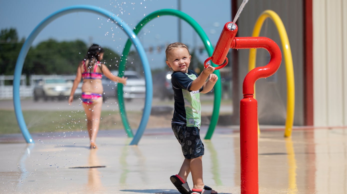 A young blonde child in a swim shirt and trunks wields a water nozzle at the CPN splash pad.