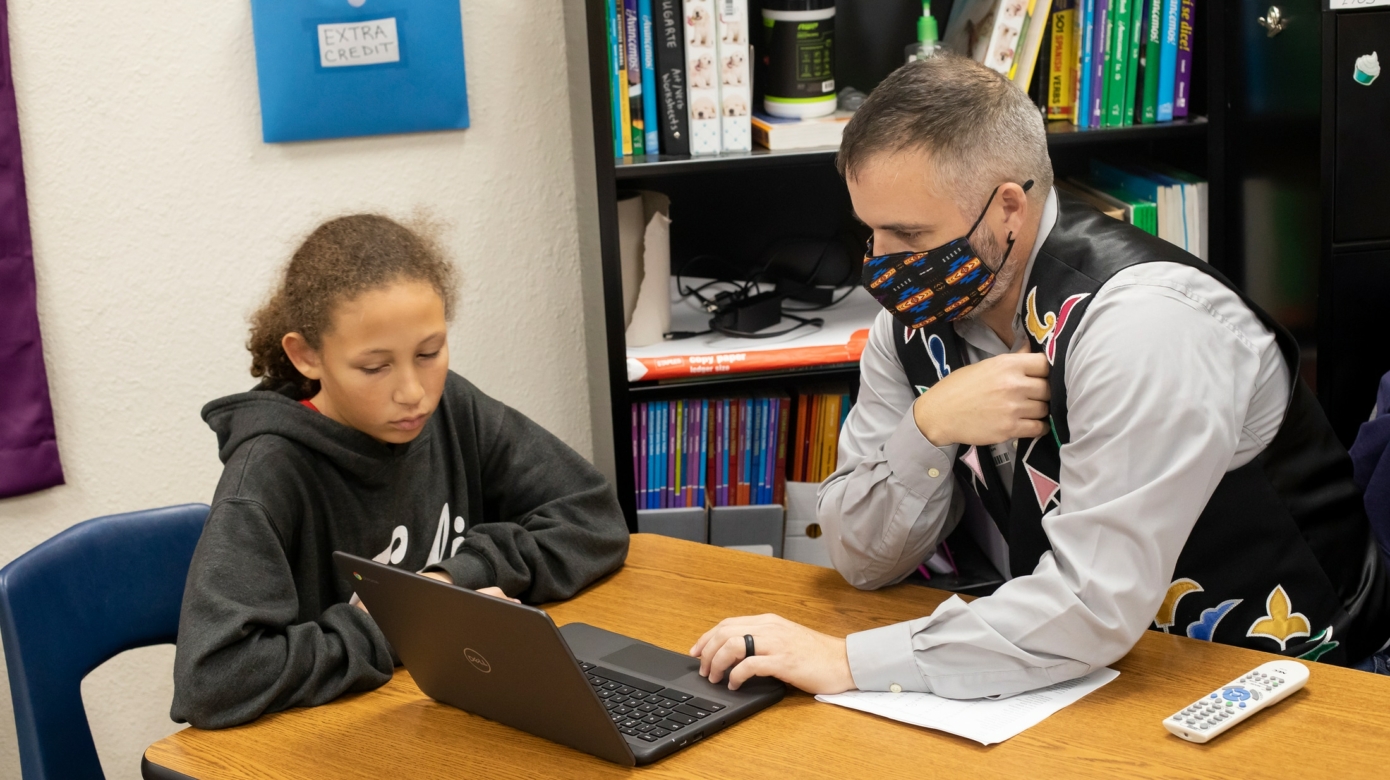 CPN Language Director Justin Neely shares a laptop with a sixth grader and Tribal member who is wearing a black sweatshirt and whose hair is pulled back into a ponytail. The two sit at a table in a Shawnee Middle School classroom.