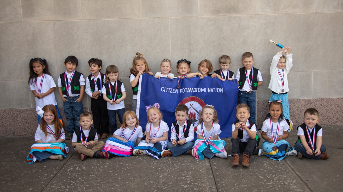 Several young children wearing matching ribbon skirts and vests hold a Citizen Potawatomi Nation flag in a group photo.