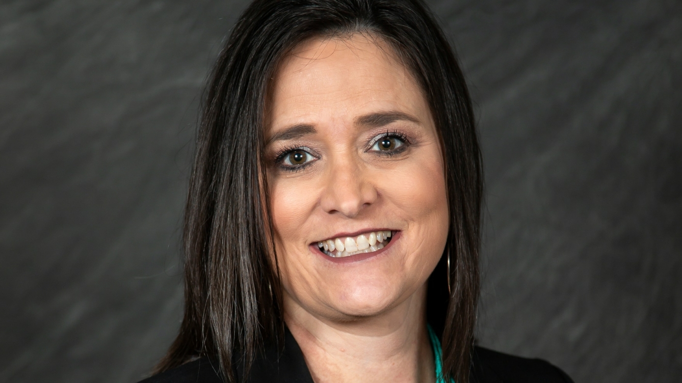 A headshot of Cindy Logsdon, CEO of CPCDC, against a dark grey backdrop. She wears a black top and a bright turquoise necklace. Her dark hair falls past her shoulders.