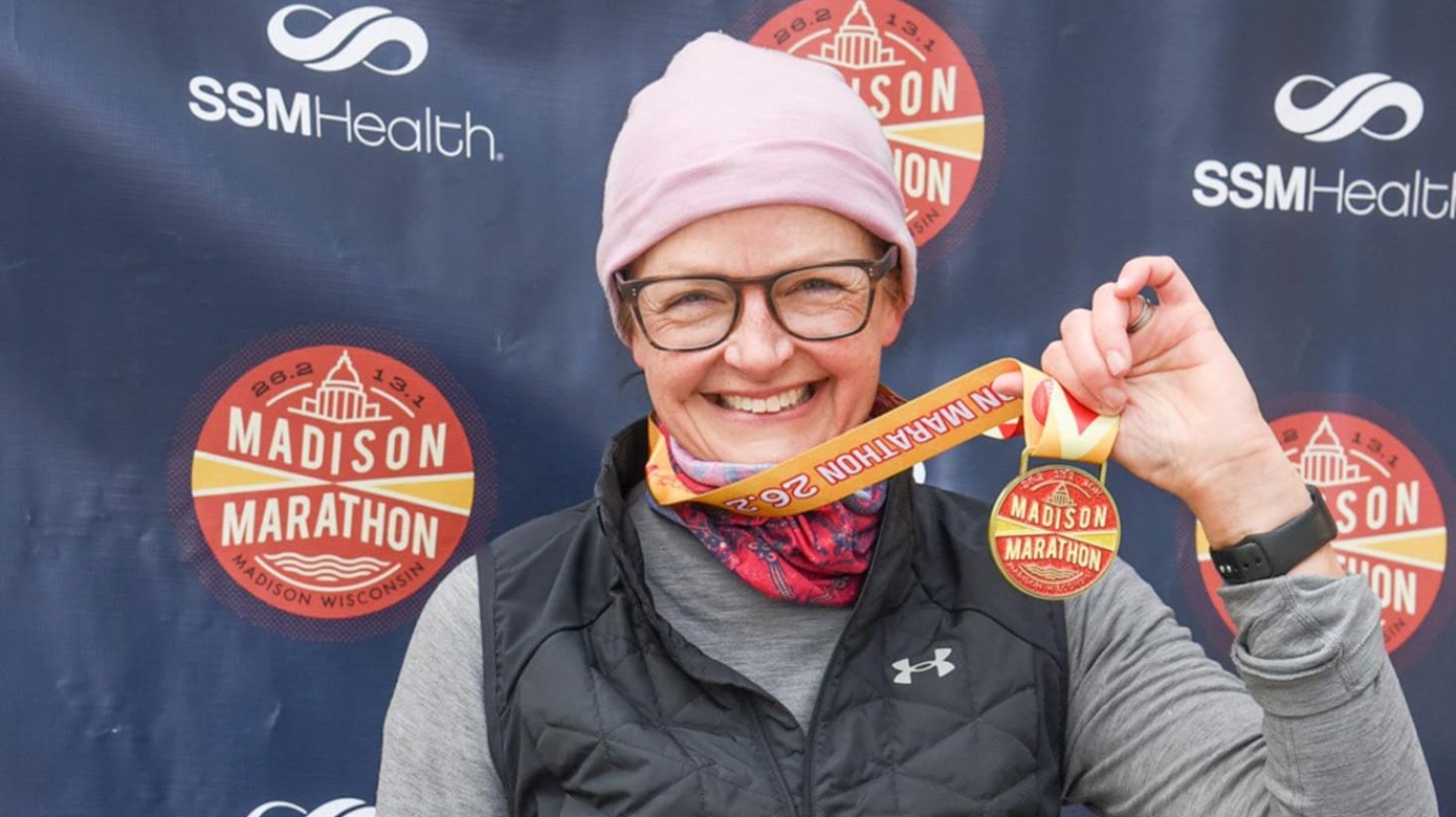 A woman wearing technical gear in greys, blues, and blacks and a light pink beanie, a yellow race bib pinned to her vest, holds up her madison Marathon finisher's medal in front of a branded marathon backdrop.