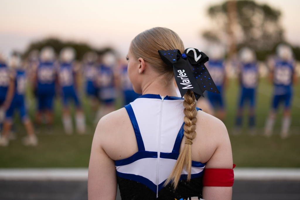 Marlee Affentranger stands with her back to the camera facing the football field. She wears a blue and white pom uniform and her blonde hair is braided in a neat ponytail with a bow bearing her name.