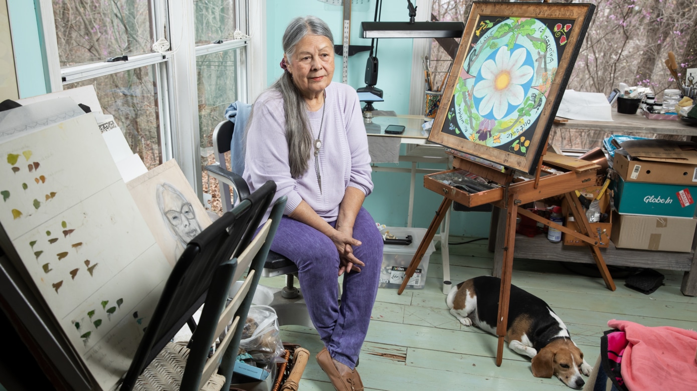 CPN Artist Minisa Crumbo Halsey, wearing shades of purple, sits in her art studio. A beagle lays under the easel, on which is round floral painting.