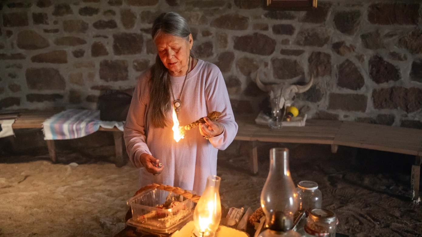 Minisa Crumbo Halsey, dressed in light purple, lights a bundle of medicine inside her longhouse.