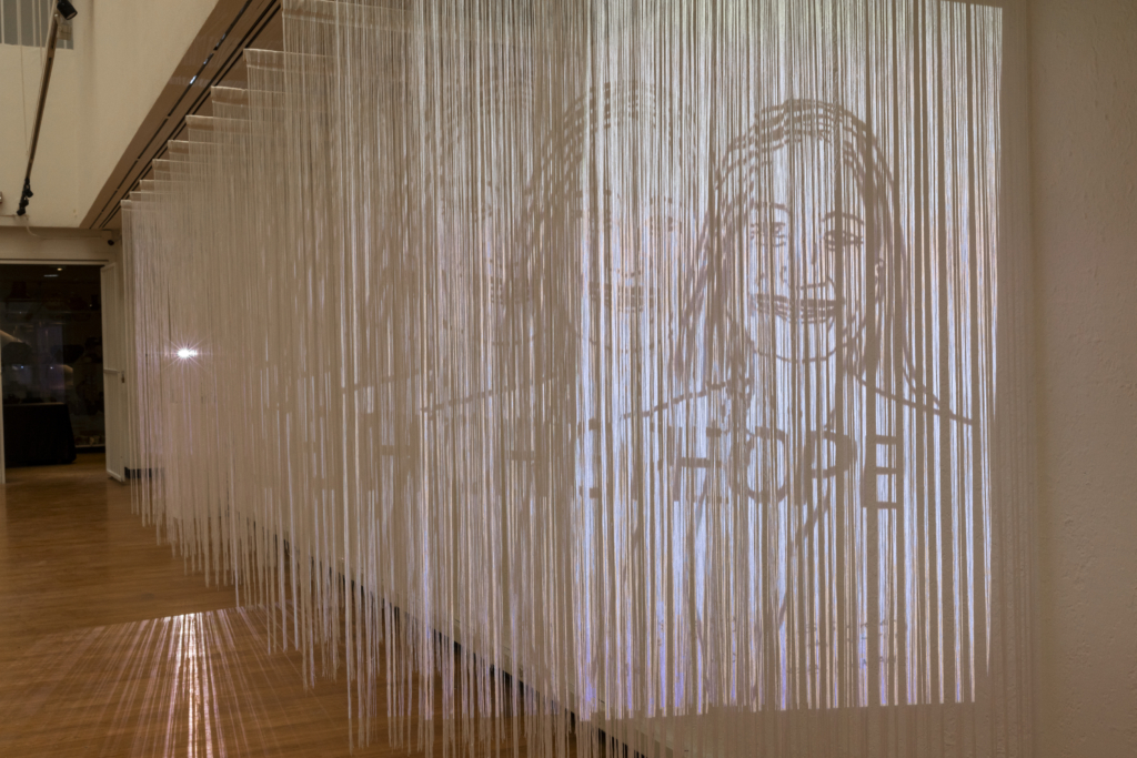Rows of white fringe hang from the ceiling. Onto the fringe is projected a drawing of a Native woman with 'HOPE' written across her chest