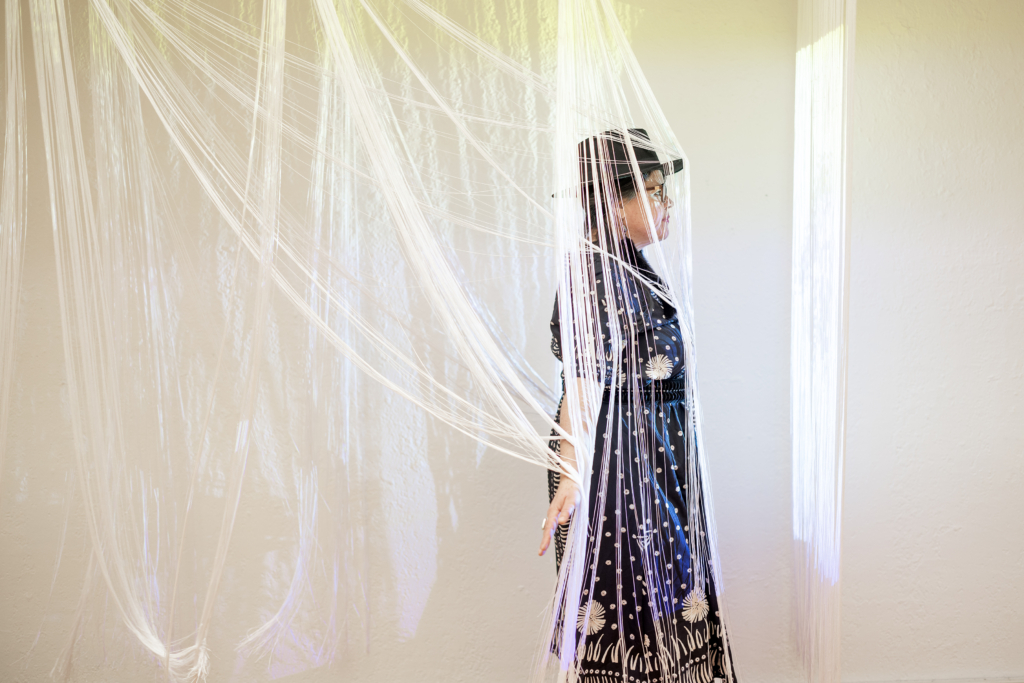 Nicole Emmons, in a black dress with white accents, walks through illuminated long white fringe hanging from the ceiling.