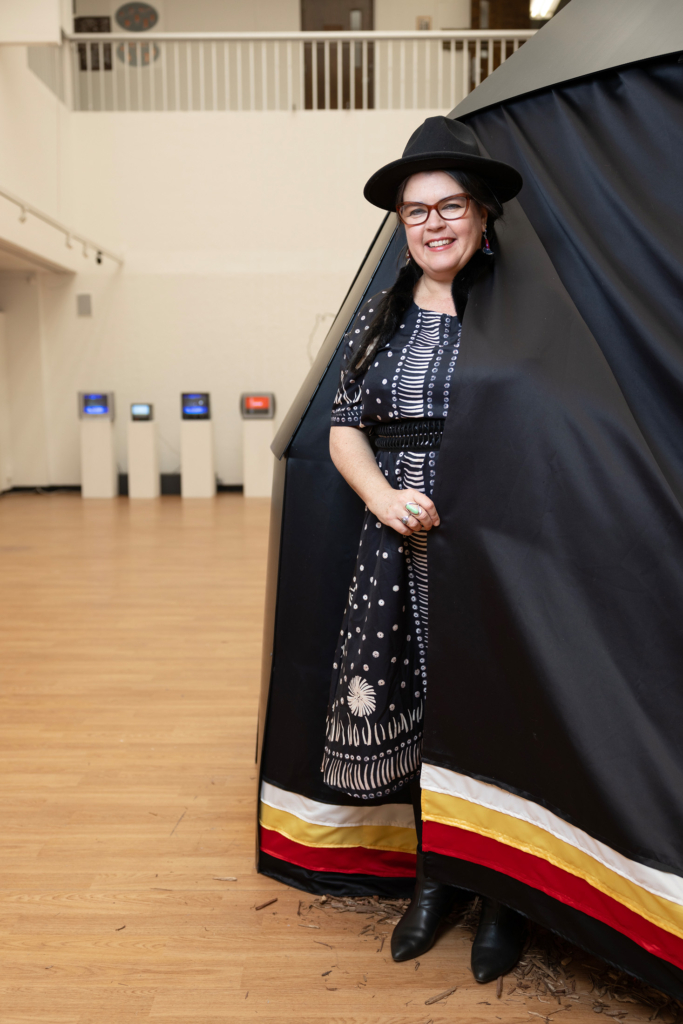 Nicole Emmons stands in the doorway of a large black wigwam structure with red, white, and yellow ribbons around the base. 