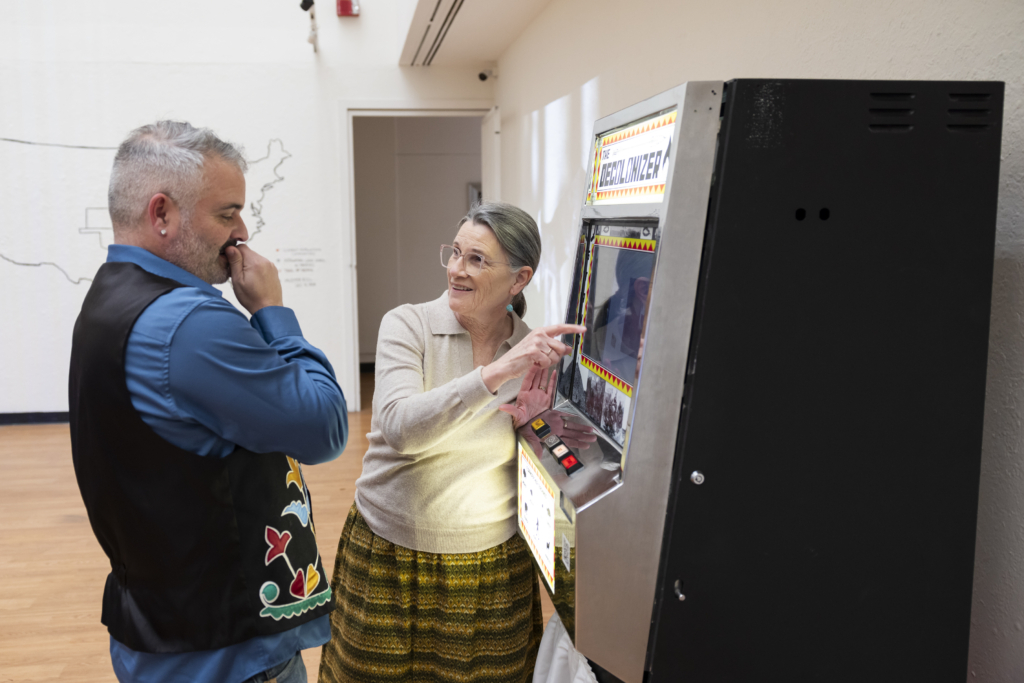 Two Tribal members examine an artistically refurbished slot machine labeled 'the Decolonizer.' 