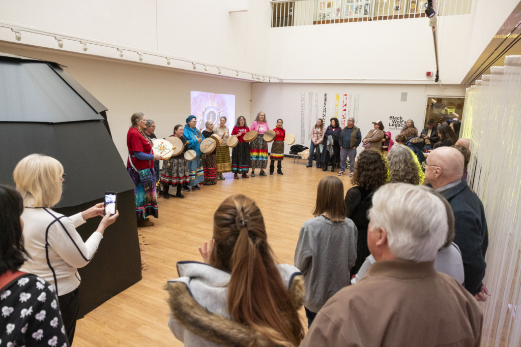 A crowd stands against one side of a gallery while 9 drummers in ribbon skirts perform along the opposite wall.