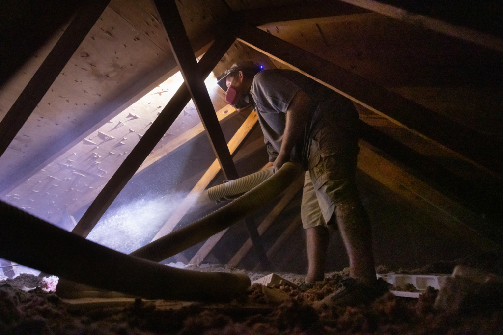 Photo of an interior of an attic where a worker is spraying insulation. The insulation is illuminated by the worker's headlamp.