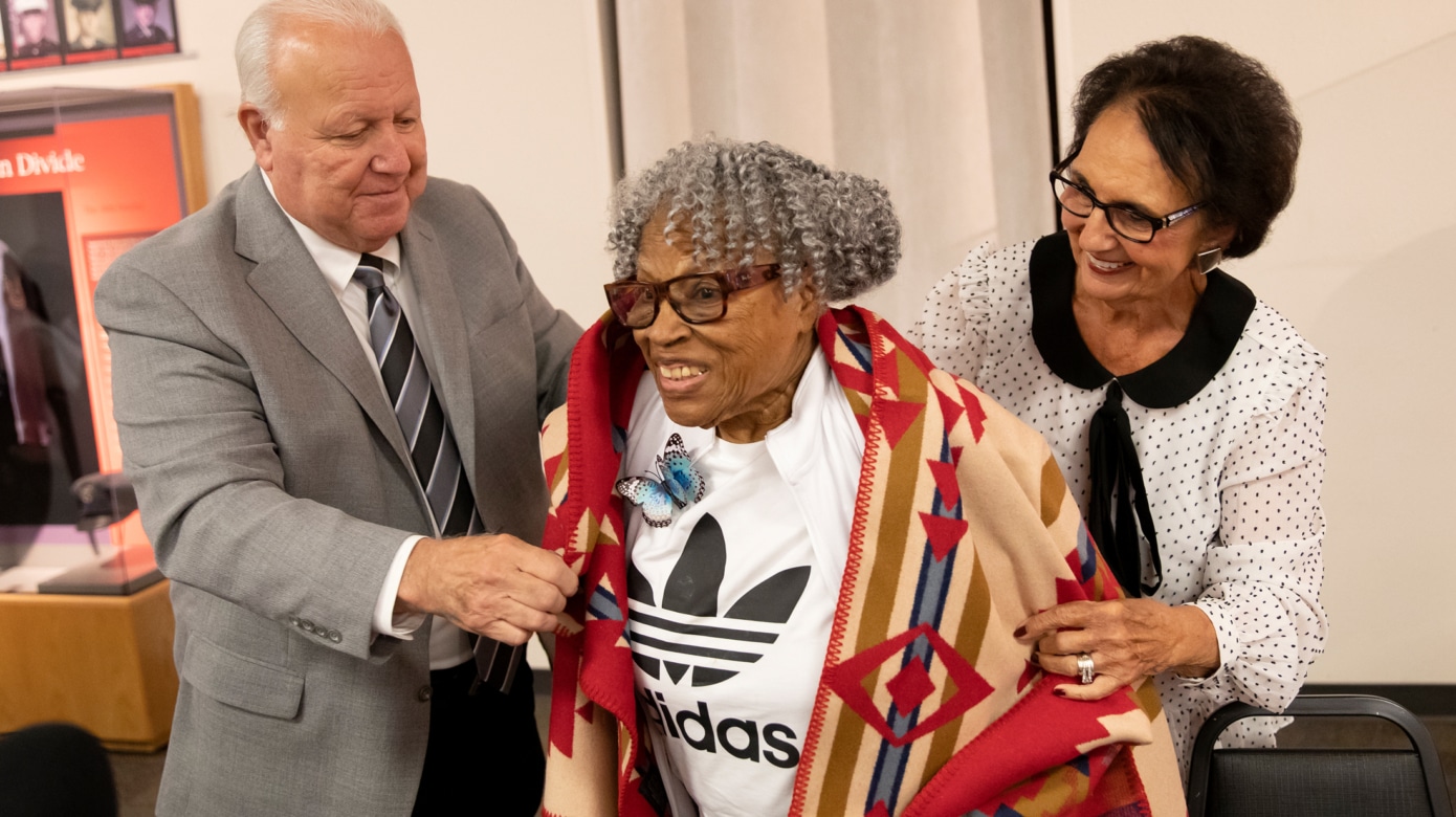 Chairman John Barrett, left, wearing a grey suit, and Vice-Chairman Linda Capps, left, wearing a black and white blouse, present Opal Lee, center, with a red and tan Pendleton blanket. Lee smiles, her grey curls fall into her face.