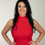 Headshot of Fire Entertainment Director and owner of DreamCatcher's Dance Company, Aoinsty Parks, in a bright red pantsuit. Her dark hair falls curled past her shoulders and she stands with hands on hips, looking at the camera.