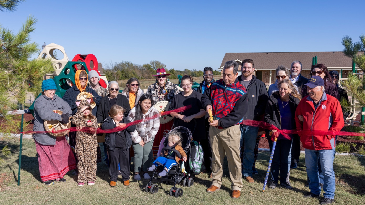 A group of people of all ages stands behind a red ribbon that Scott George, front and center, cuts.