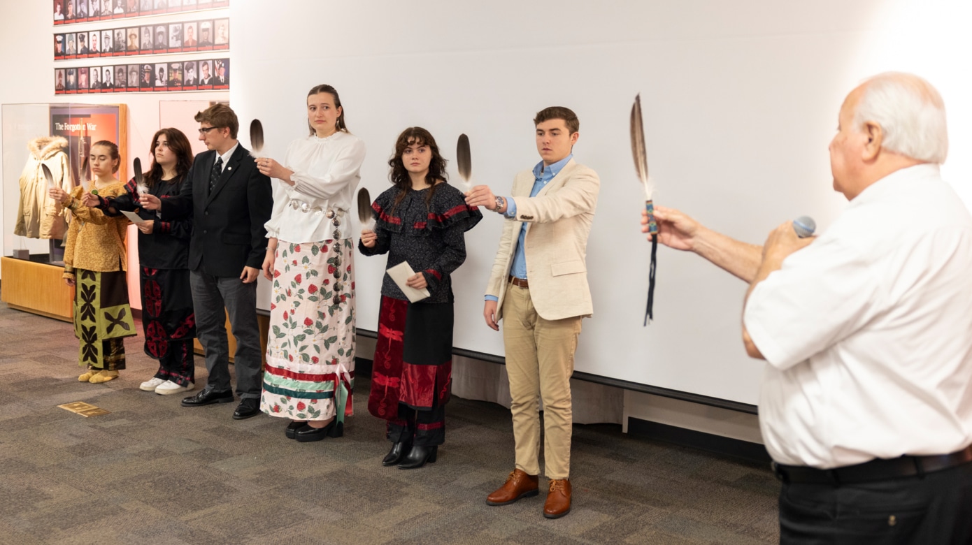 Six students in regalia hold up eagle feathers in front of them. CPN Chairman Barrett, far right, demonstrates for the students.
