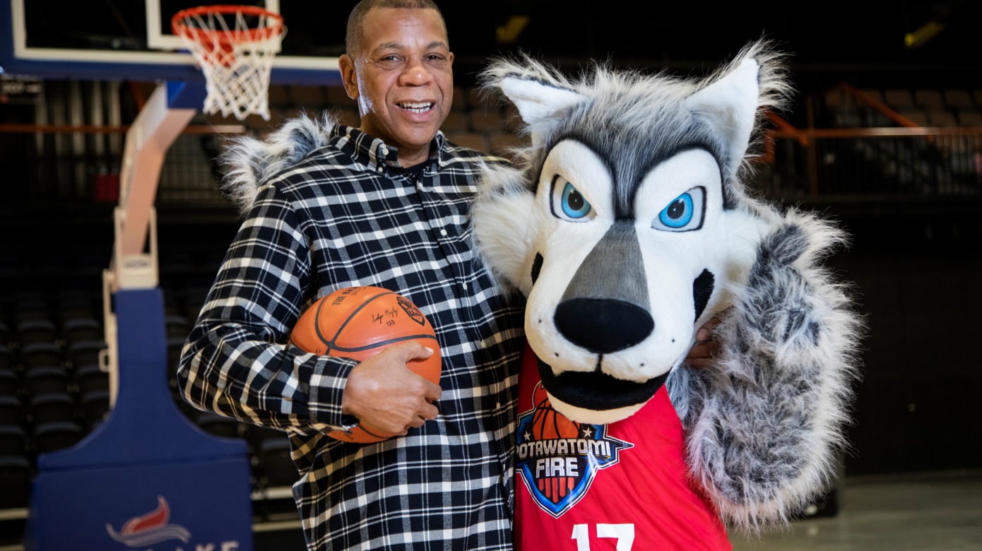 Potawatomi Fire head coach Derrick Rowland, wearing a navy plaid shirt and holding a basketball in his right hand, poses with the Fire's new mascot, a wolf named Mo.