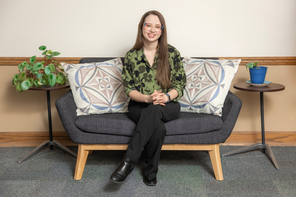 Portrait of Rachel Watson, wearing a green floral blouse and black pants, seated on a couch in the Department of Education office.