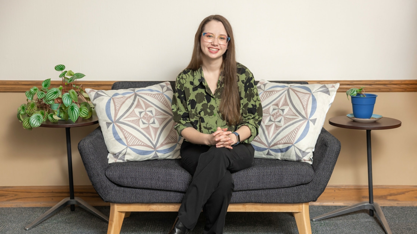 Portrait of Rachel Watson, wearing a green floral blouse and black pants, seated on a couch in the Department of Education office.