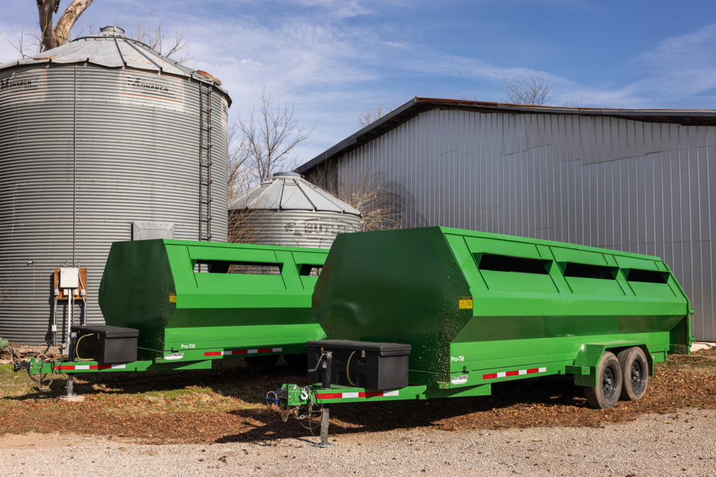 Two large Deere green trailers pictured in front of a metal barn and silo.