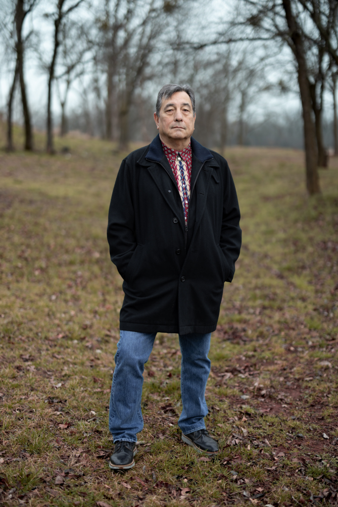 Scott George, wearing a red patterened shirt visible beneath a pea coat and zip sweater, looks solemly into the camera in an outdoor portrait.