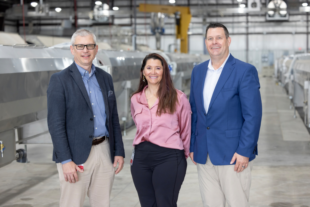 Jeremy Hohn, wearing khaki pants, a blue shirt, and navy blazer; Barb Donaldson, wearing black slacks and a pink blouse; and Ronnie Wear, wearing khaki pants, a white shirt, and blue blazer, pose inside Sovereign Pipe Technologies' warehouse.