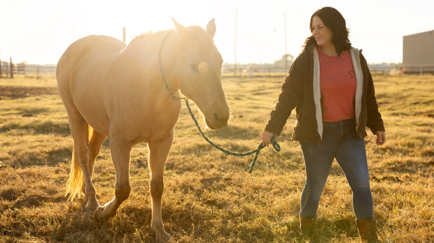 Tribal member Jessica King, wearing jeans, a pink t-shirt, and a zip-up jacket, leads a brown horse through a field at golden hour.