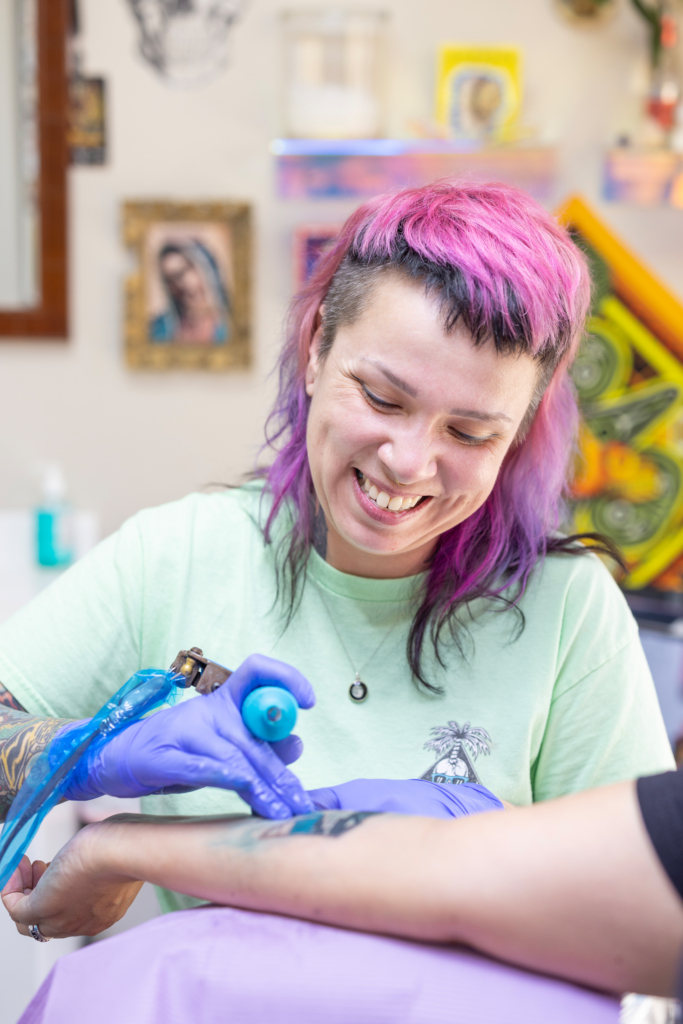 Shelly Wahweotten smiles while working on a tattoo on a client's forearm. Wahweotten's hair is dyed pink and purple, and she wears a green t-shirt. 