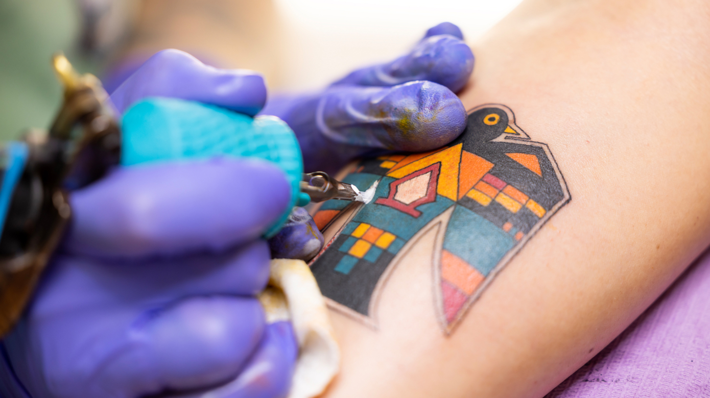Close shot of a tattoo artist's hands in purple gloves putting final touches on a colorful thunderbird medallion tattoo.