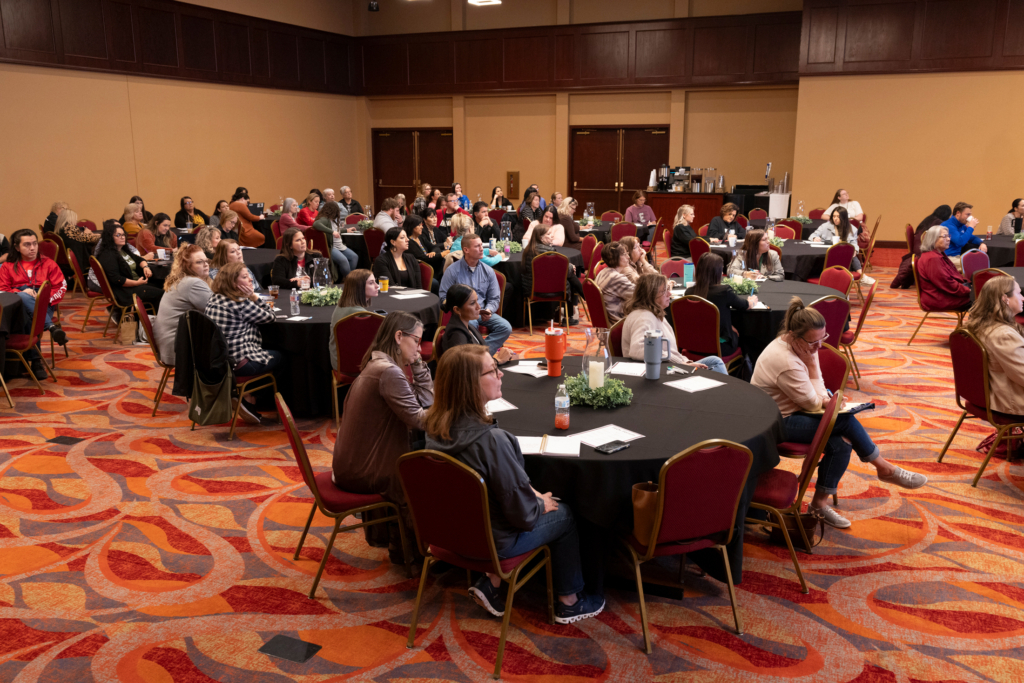 Long shot of a room full of round tables covered with black table cloths. Attendees sit around the tables, listening to speakers at the front of the room.