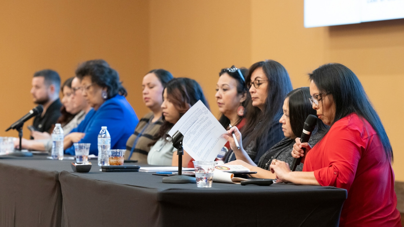 A row of panelists sit at a table covered with a black table cloth. Assorted microphones, papers, and water bottles are also on the table.