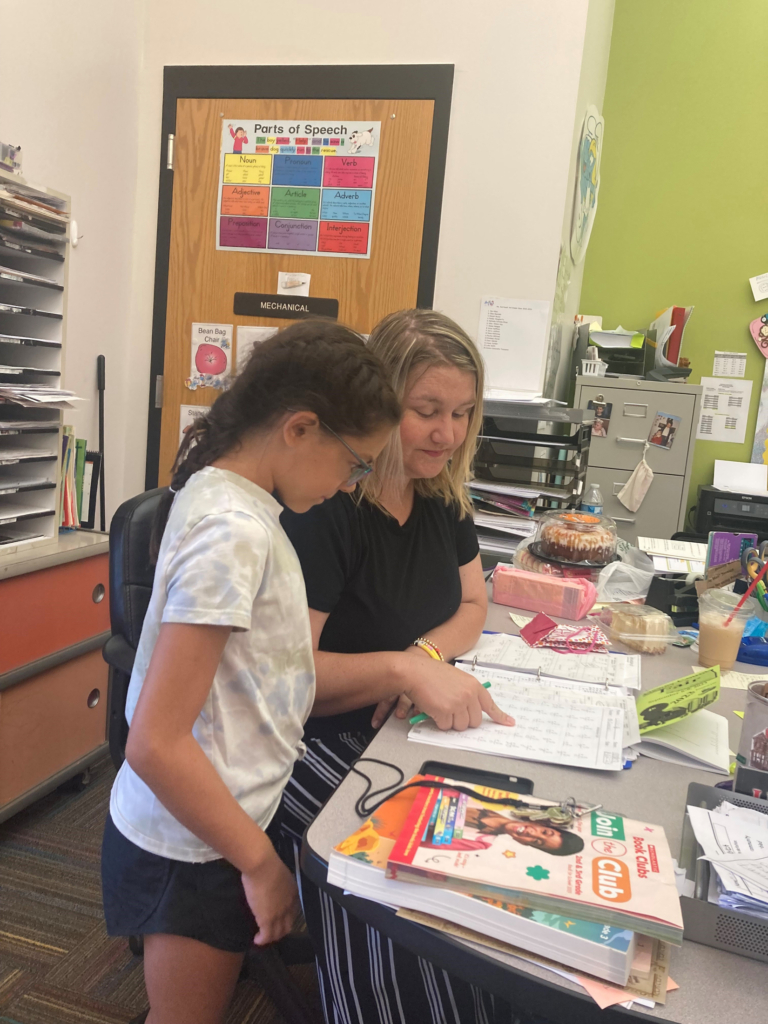 Photograph of teacher Sarah Garrison seated at a desk covered with worksheets and books, looking at a worksheet with a young student who stands next to her.