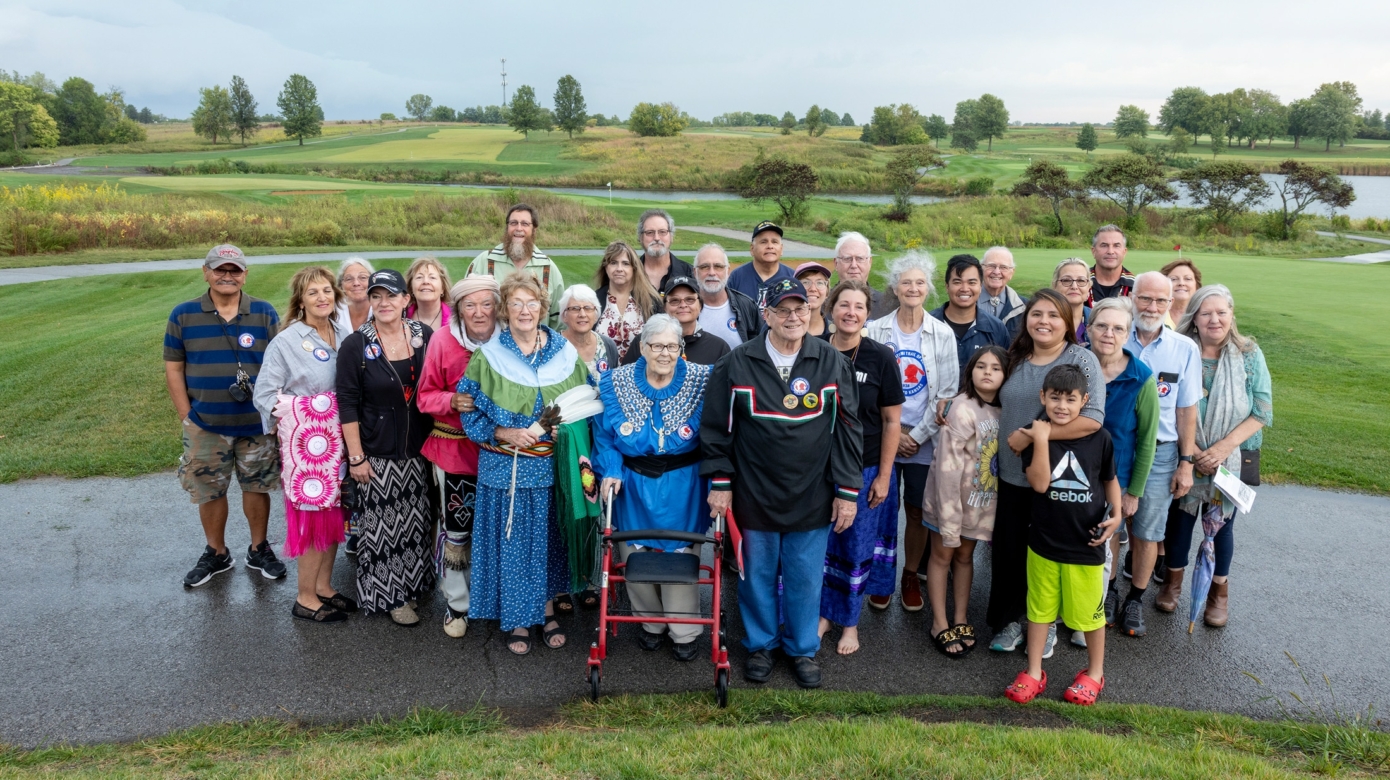 Photo of a large group standing on a paved pathway with green fields and trees behind them on a drizzly day.