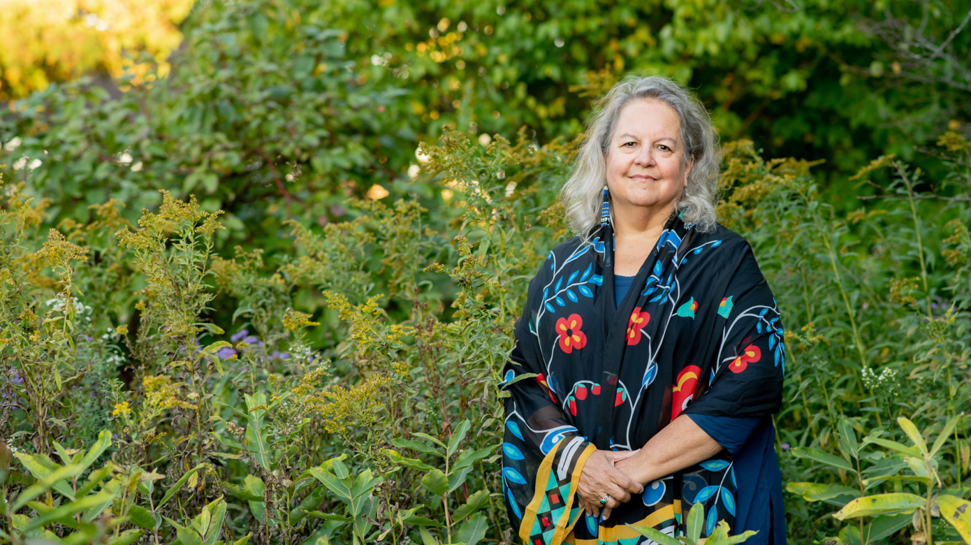Dr. Robin Wall Kimmerer stands among vibrant green plants reaching to her shoulders. She wears a black shawl with brightly colored floral patterns on it.