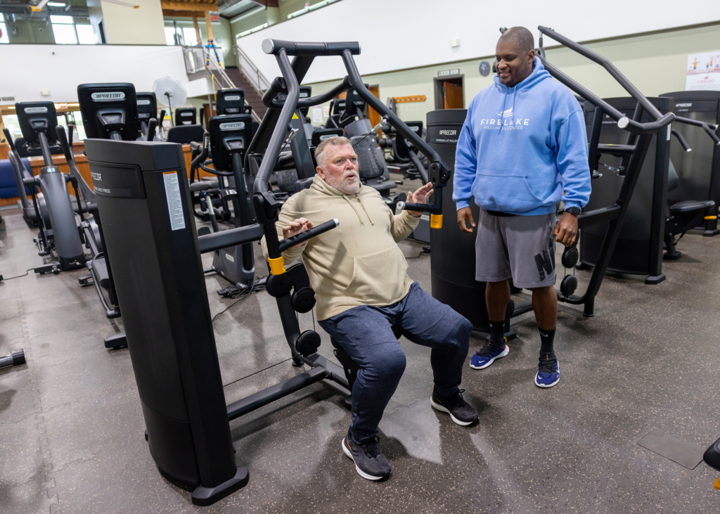 A FireLake Wellness Center staff member wearing a light blue Wellness Center hoodie and grey Nike shorts looks on as a client in a brown sweatshirt and blue sweatpants utilizes a weight machine.