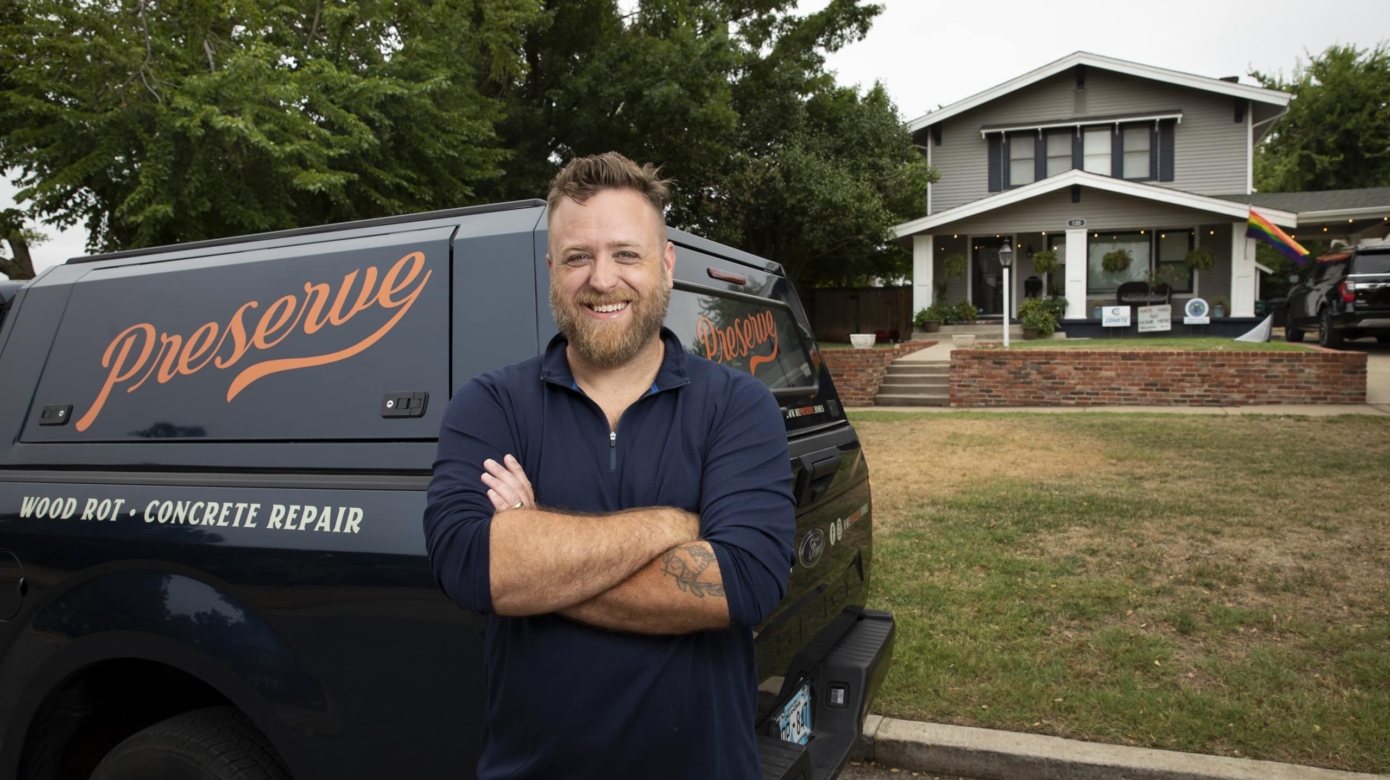 Ty McBride stands with his arms folded across his navy quarter zip and smiles at the camera in front of his We Preserve Homes van and a house he worked on.