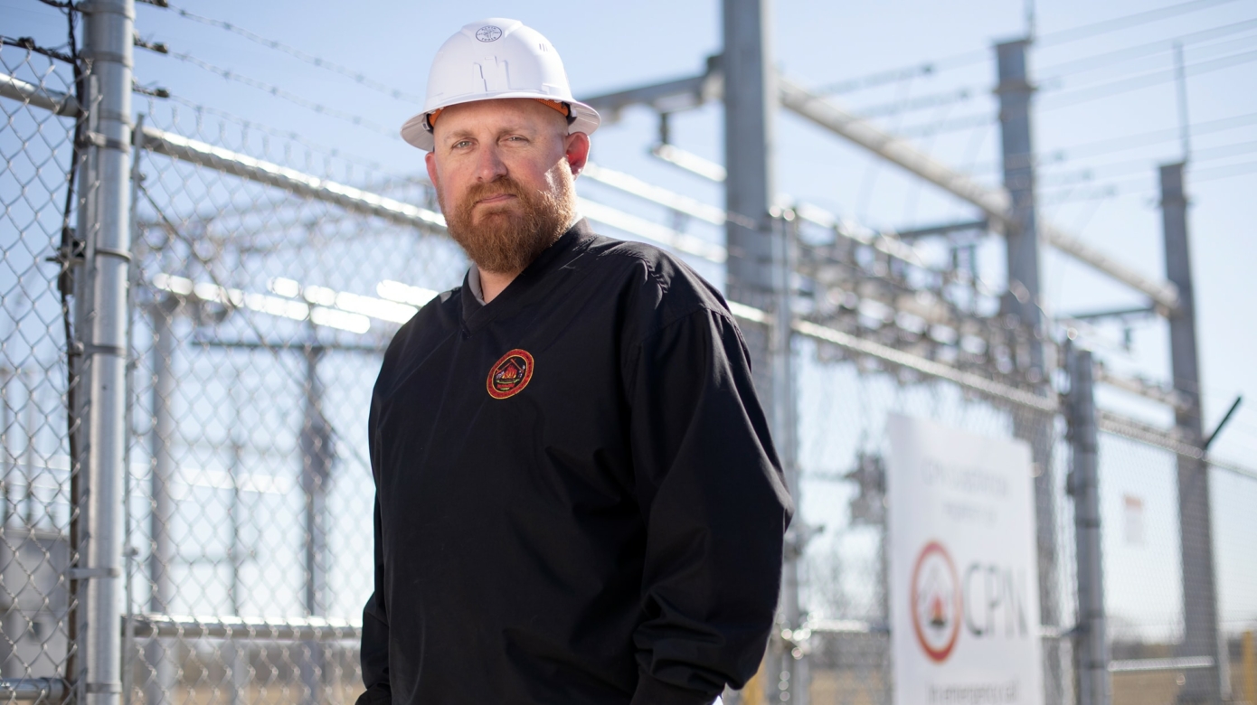 Portrait of CPN Electrical Director Justin Whitecotton at CPN's substation. Lines of a chain-link fence, electrical wires and poles criss-cross the background of the photo.