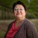 Headshot of CPN Workforce and Social Services Director Margaret Zientek. She wears muted red lipstick and frameless glasses, and stands in front of trees which are out of focus in the background.