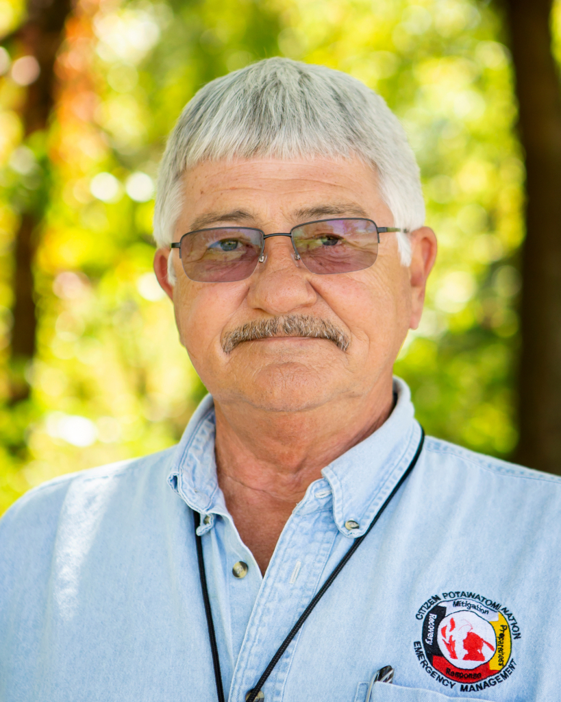 A portrait of Tim Zientek, with grey hair and mustache, rectangular transition lens glasses slightly darkened, and a CPN Emergency Services button-up shirt in front of bright green tree leaves blurred from focus in the background.